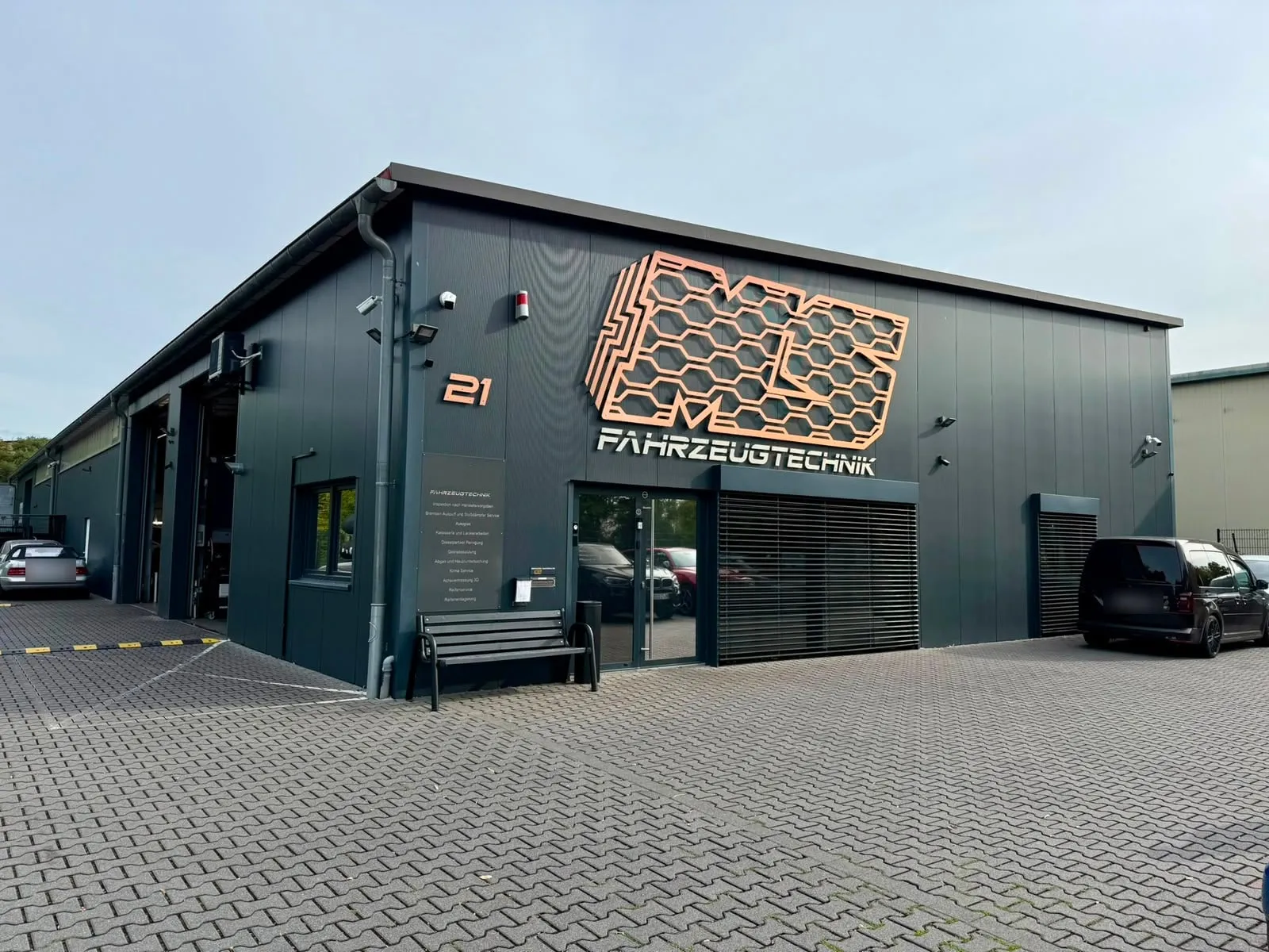 Betriebsgebäude der MS Fahrzeugtechnik in Maintal-Dörnigheim: dunkel verkleidete Halle mit bronzefarbenem Wabenlogo über dem Eingang, davor der gepflasterte Hof mit den Kundenparkplätzen